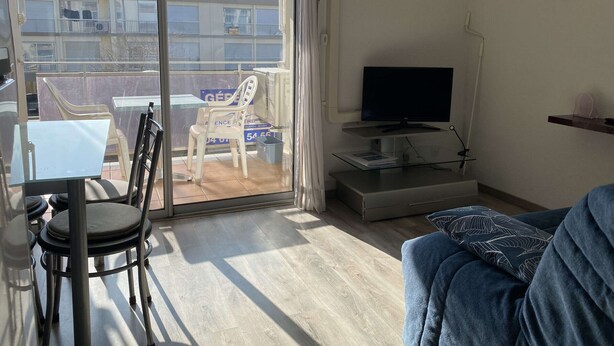 A living room with a TV, a sofa, a dining table with chairs, and a balcony with a chair and a table.
