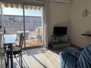 Een woonkamer met een televisie, een bank, een eettafel met stoelen en een balkon met een stoel en een tafel.