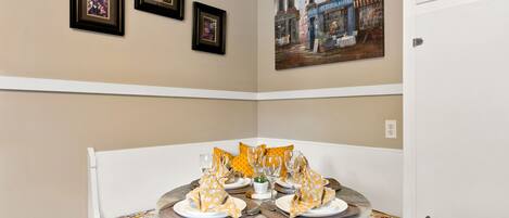 A dining area with a round table set for four, framed pictures on the wall, and a painting depicting a street scene.