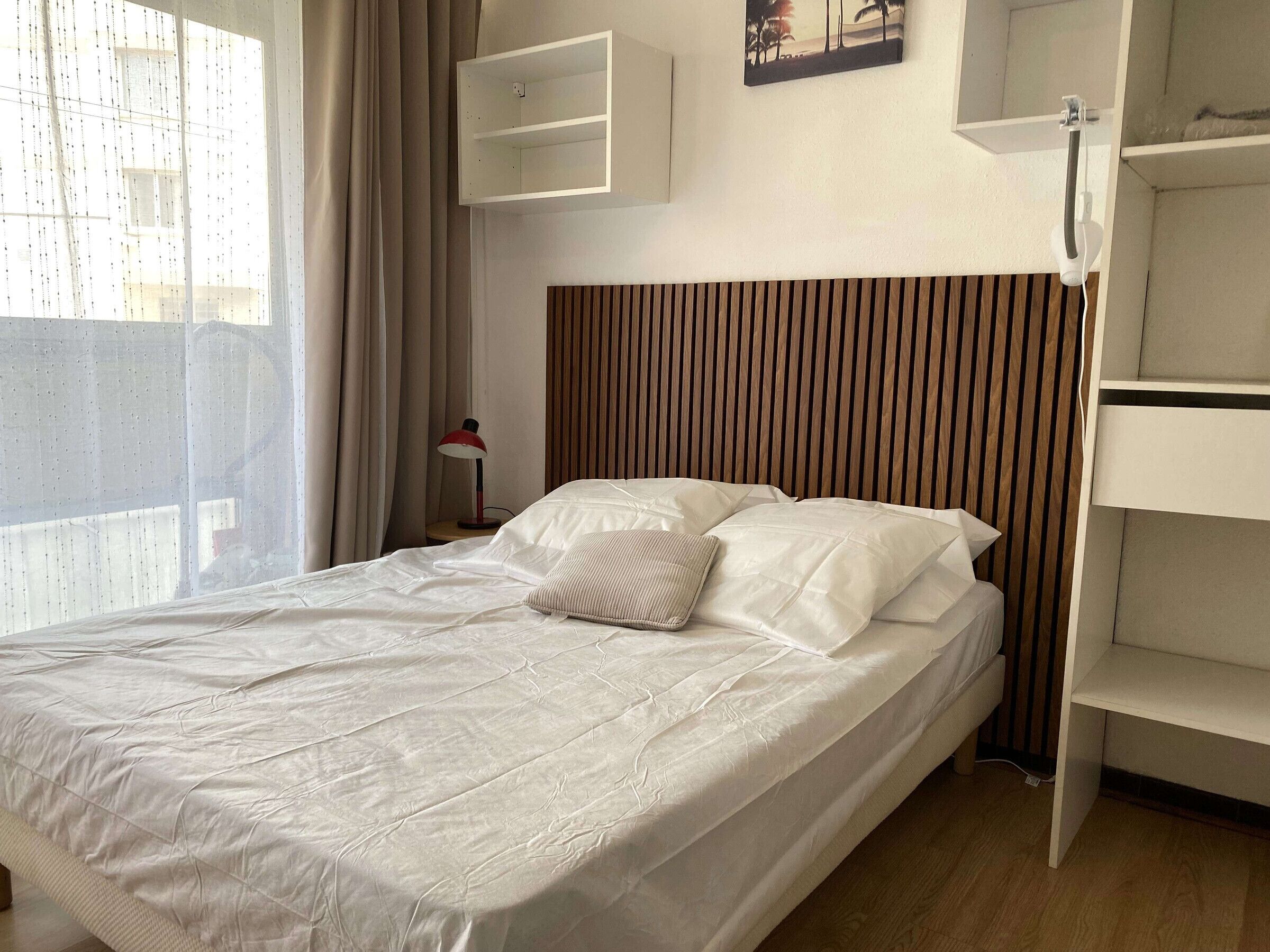 A neatly made bed with white linens, a wooden slatted headboard, a bedside table with a lamp, and a wall-mounted shelf.