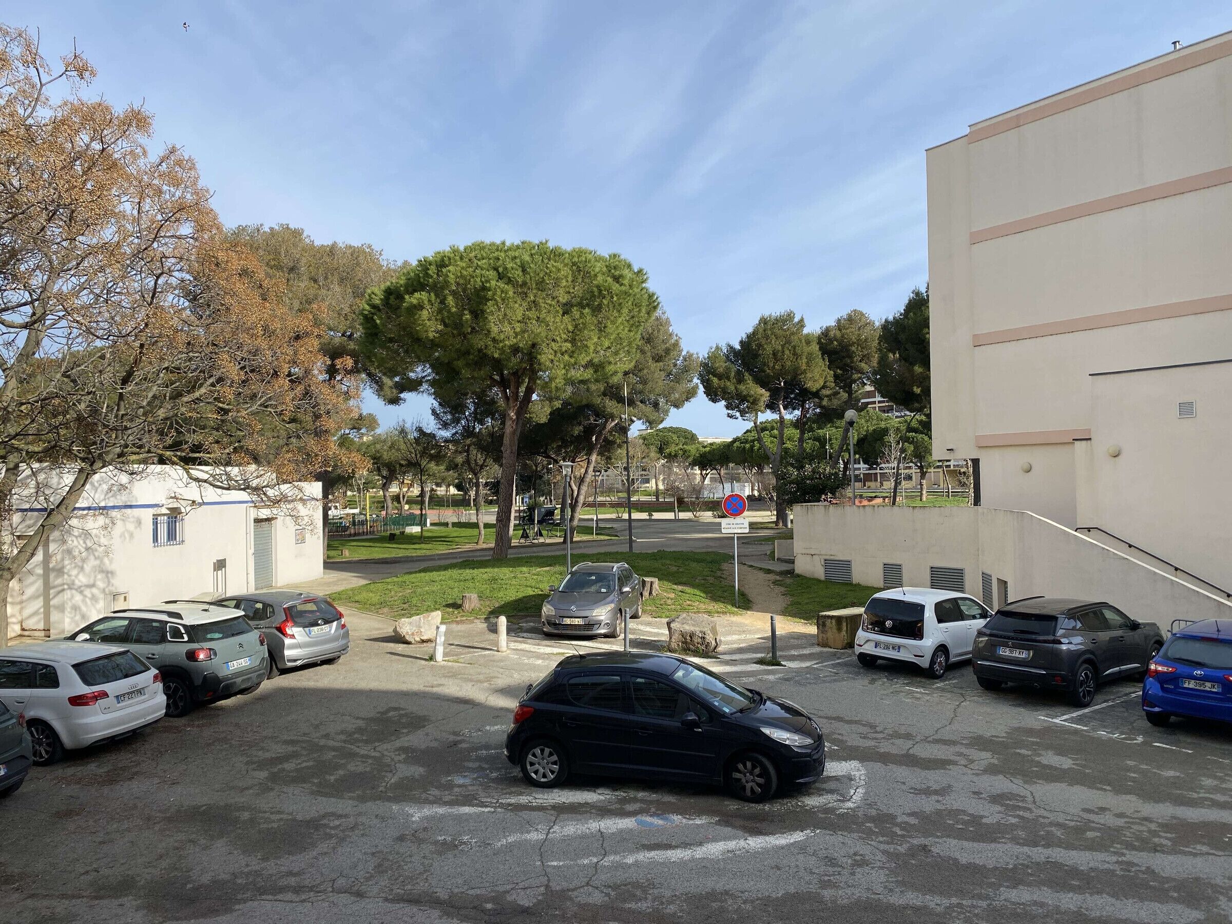 A parking area with multiple cars, a building with a beige facade, and a tree-lined street.