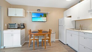 A compact kitchen with white cabinets, a wooden dining table, and a TV displaying a beach scene.
