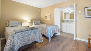 A bedroom with two beds, a nightstand, a lamp, a painting, and a bathroom visible through an open door.