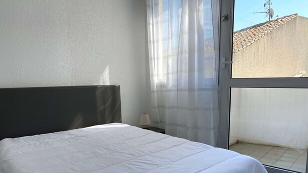 Een bed met een witte dekbedovertrek, een donker hoofdbord, een bedlampje en een uitzicht op een gebouw door een raam met dunne gordijnen.