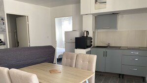 Een kleine keuken met een spoelbak, kookplaat en kastjes. Er is een eethoek met een tafel en stoelen.
