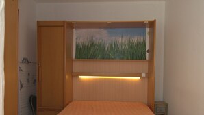 A small bedroom with a bed, a wooden wardrobe, a nightstand, and a wall-mounted shelf with a painting. - Cozy T1 in Balaruc-les-Bains with Balcony, A/C, and Modern Amenities (Balaruc-les-Bains)