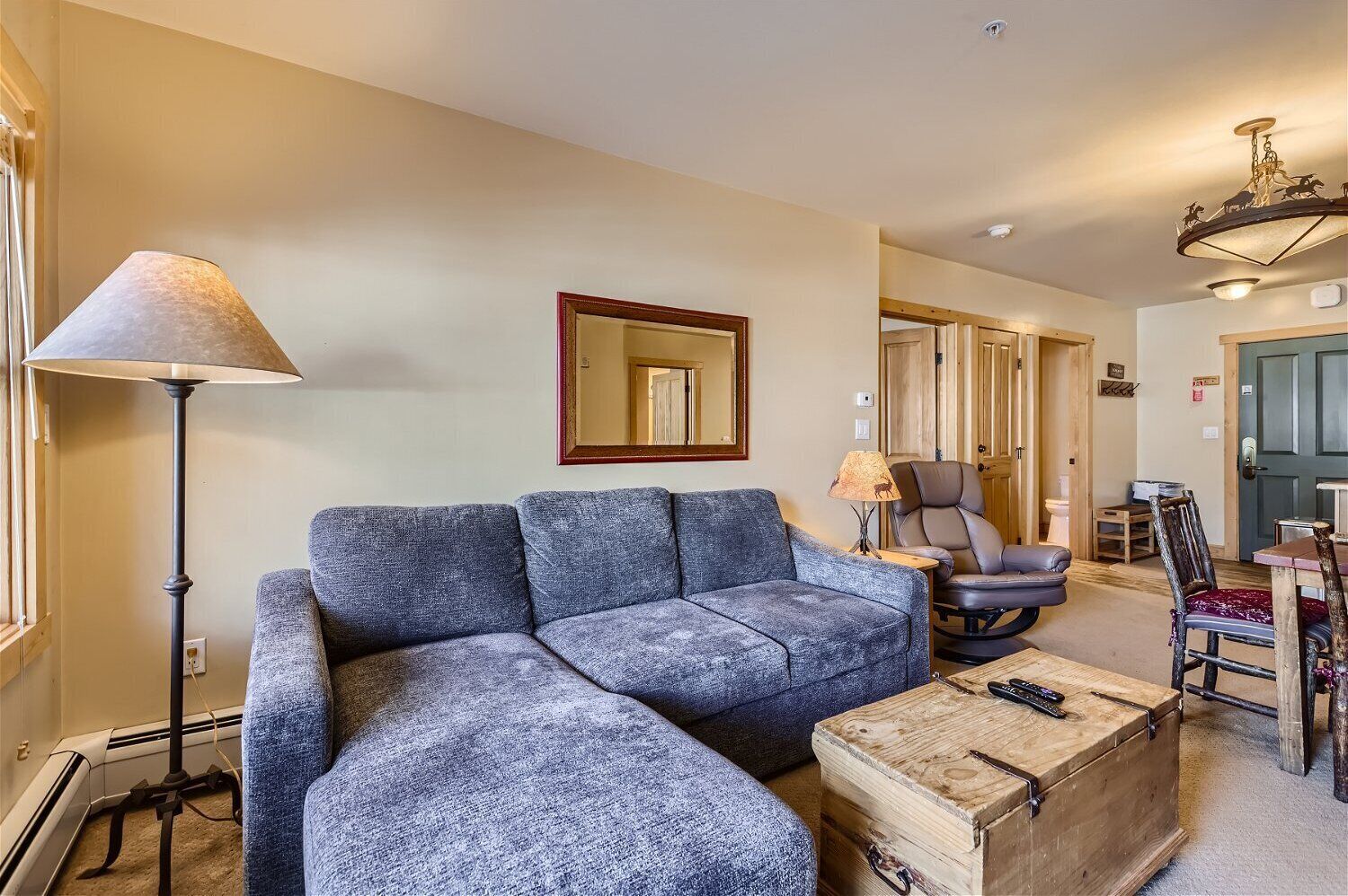 A living room with a sectional sofa, a floor lamp, a wooden chest, and a dining area with chairs and a table.