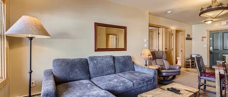A living room with a sectional sofa, a floor lamp, a wooden chest, and a dining area with chairs and a table.