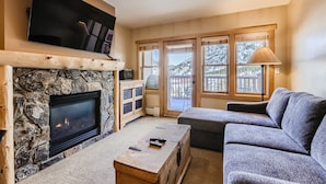 A cozy living room with a stone fireplace, a flat-screen TV mounted above it, and a large window overlooking a deck. - Updated 2 Bedroom Walking Distance to the SuperBee Lift!! (Frisco)