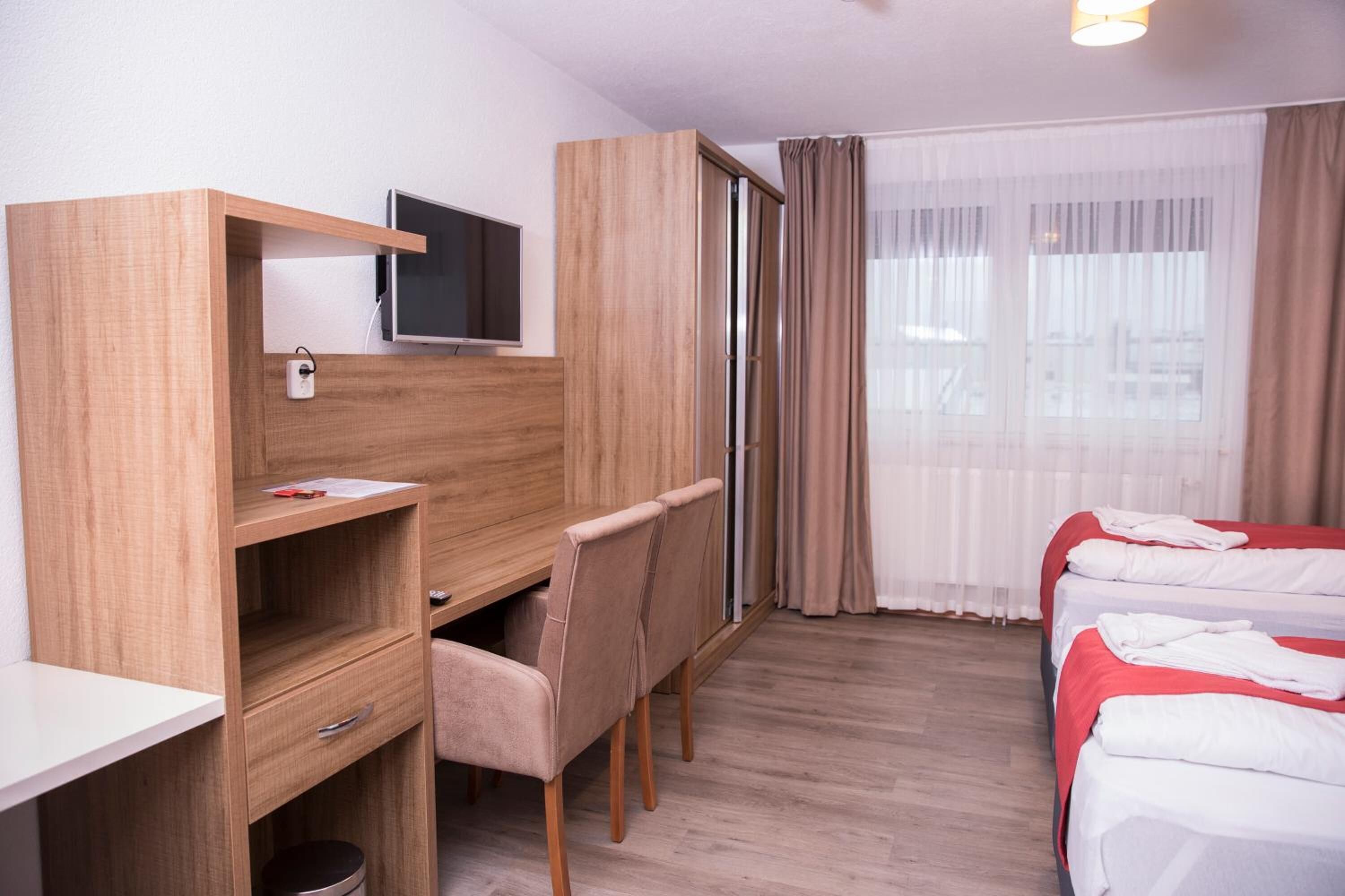A hotel room with a wooden desk, a television, a chair, a bed with red bedding, and a window with curtains.
