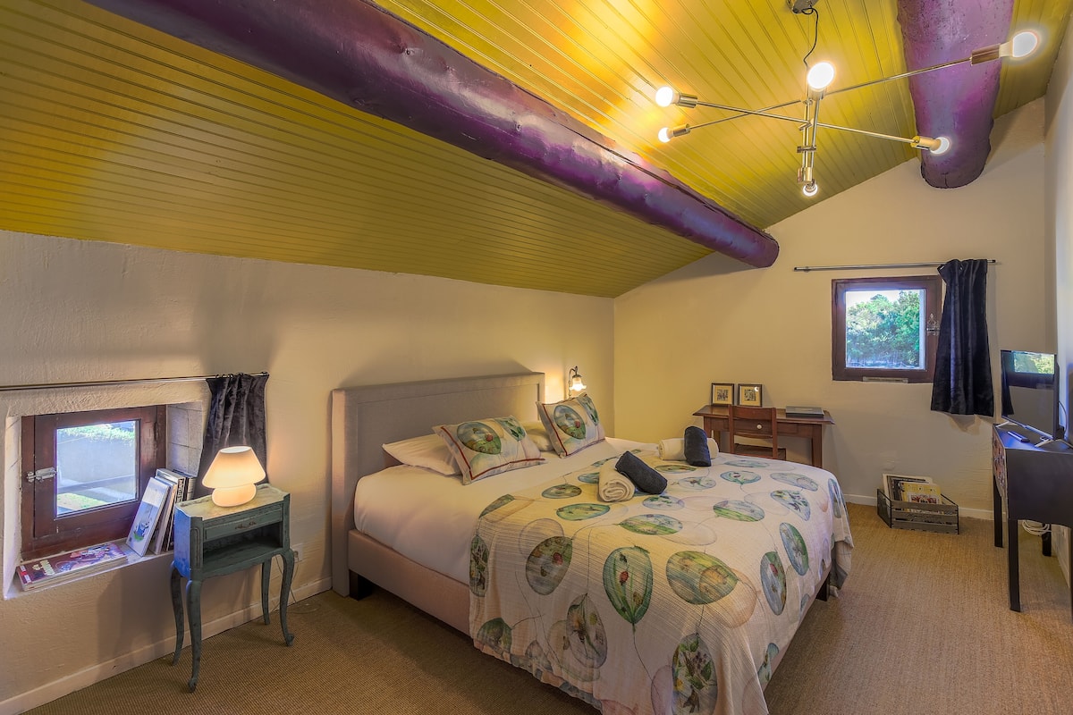 A bedroom with a bed, bedside table, lamp, window, and a desk with a chair.