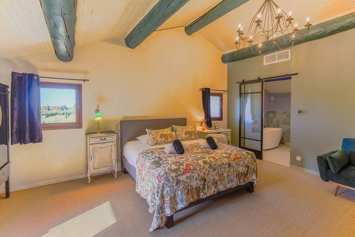 A bedroom with a large bed, a nightstand, a lamp, a chair, a window with curtains, and a bathroom visible through a glass door.