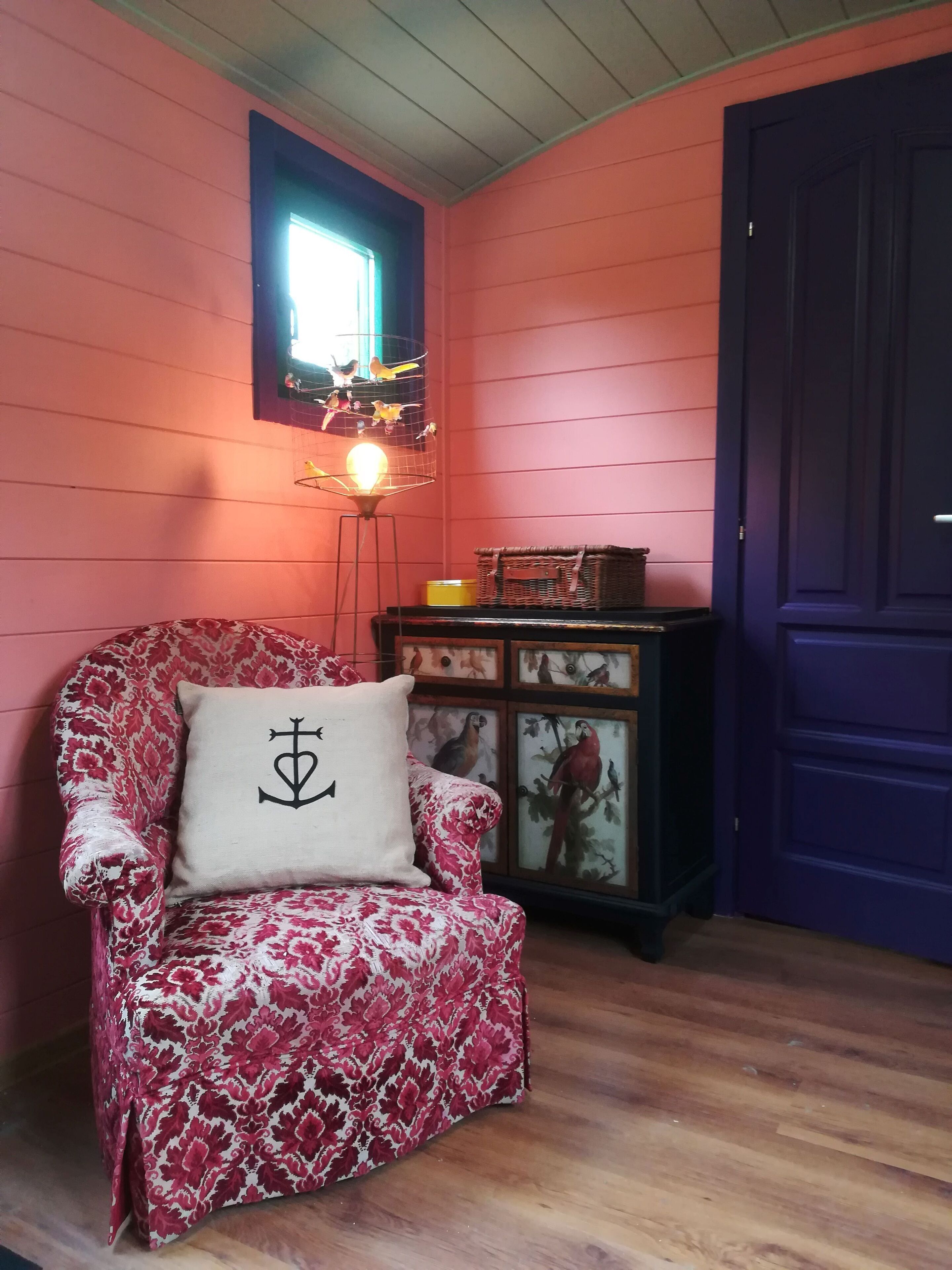 A room with a red wall, a wooden floor, a chair with a floral pattern, a cushion with a symbol, a lamp, a small table with a basket, and a dark blue door.
