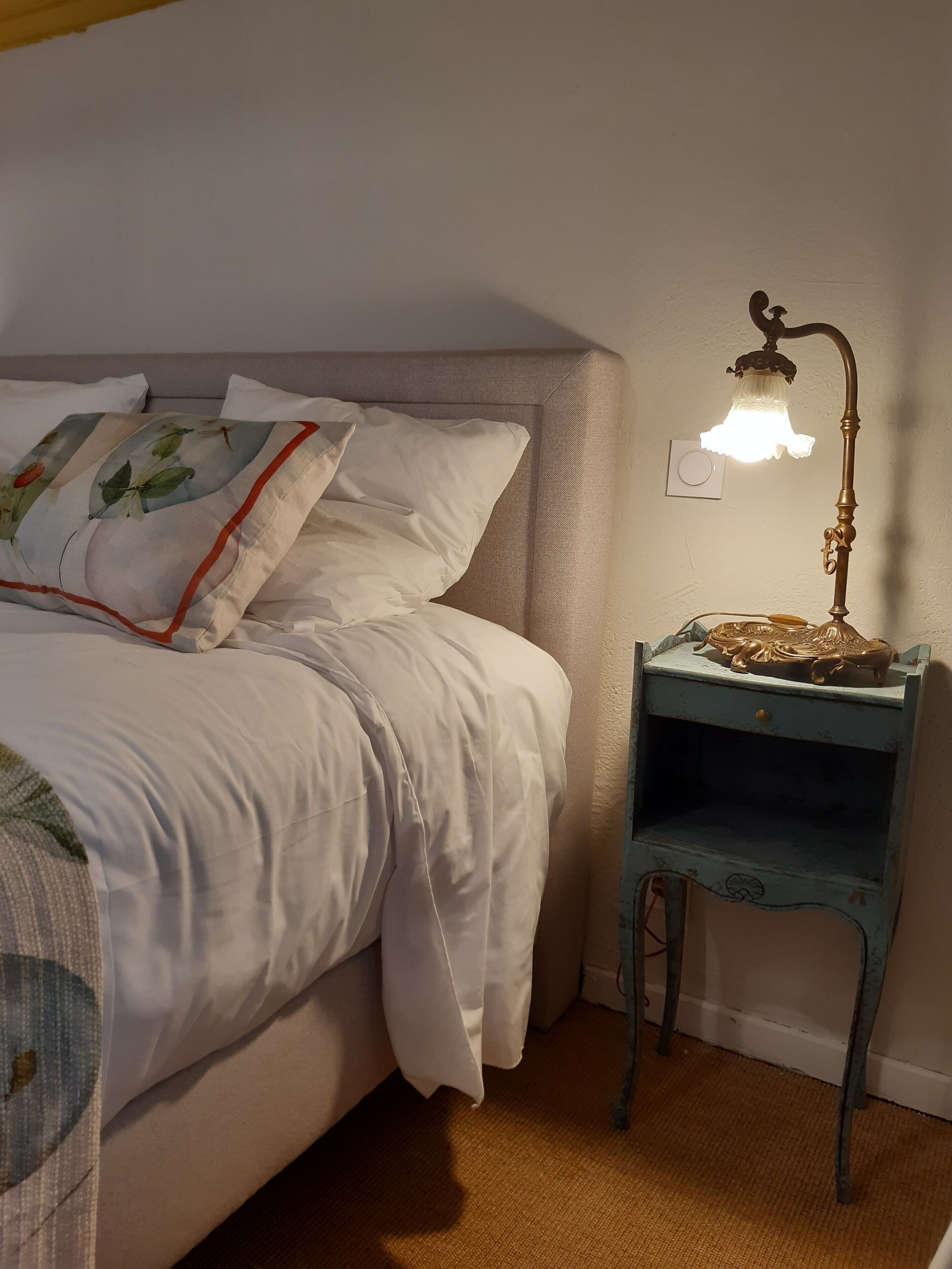 Ein ordentlich bezogenes Bett mit weißen Leinen und einem Blumenkissen, ein kleiner Nachttisch mit Lampe und eine Steckdose.