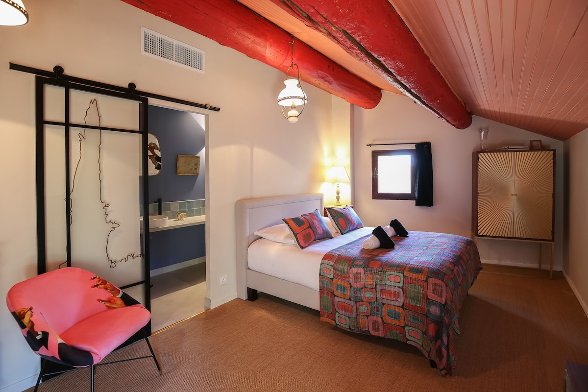 A bedroom with a bed, a chair, a window, and a wall with a decorative piece.