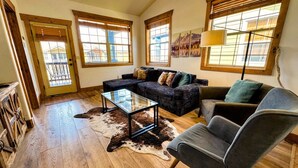 A living room with a sectional sofa, a glass coffee table, a leather armchair, wooden flooring, and large windows. - Pet-Friendly Driggs Condo Near Targhee (Driggs)