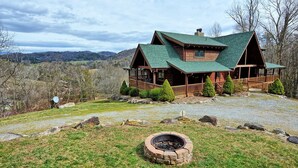 A log cabin with a green roof, a porch, and a fire pit in the foreground. - Experience luxury in this 6-bedroom cabin in Sugar Grove, NC, ideal for 12! (Sugar Grove)