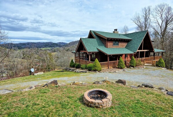 A log cabin with a green roof, a porch, and a fire pit in the foreground.