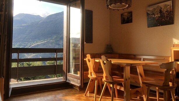 A dining area with a wooden table and chairs, a large window with a view of mountains, and a hanging light fixture. - Spacious, 6-person chalet with stunning views in La Norma, French Alps (La Norma)