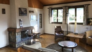 A cozy living room with a stone fireplace, wicker furniture, and a large window with curtains. - Spacious, 6-person chalet with stunning views in La Norma, French Alps (La Norma)