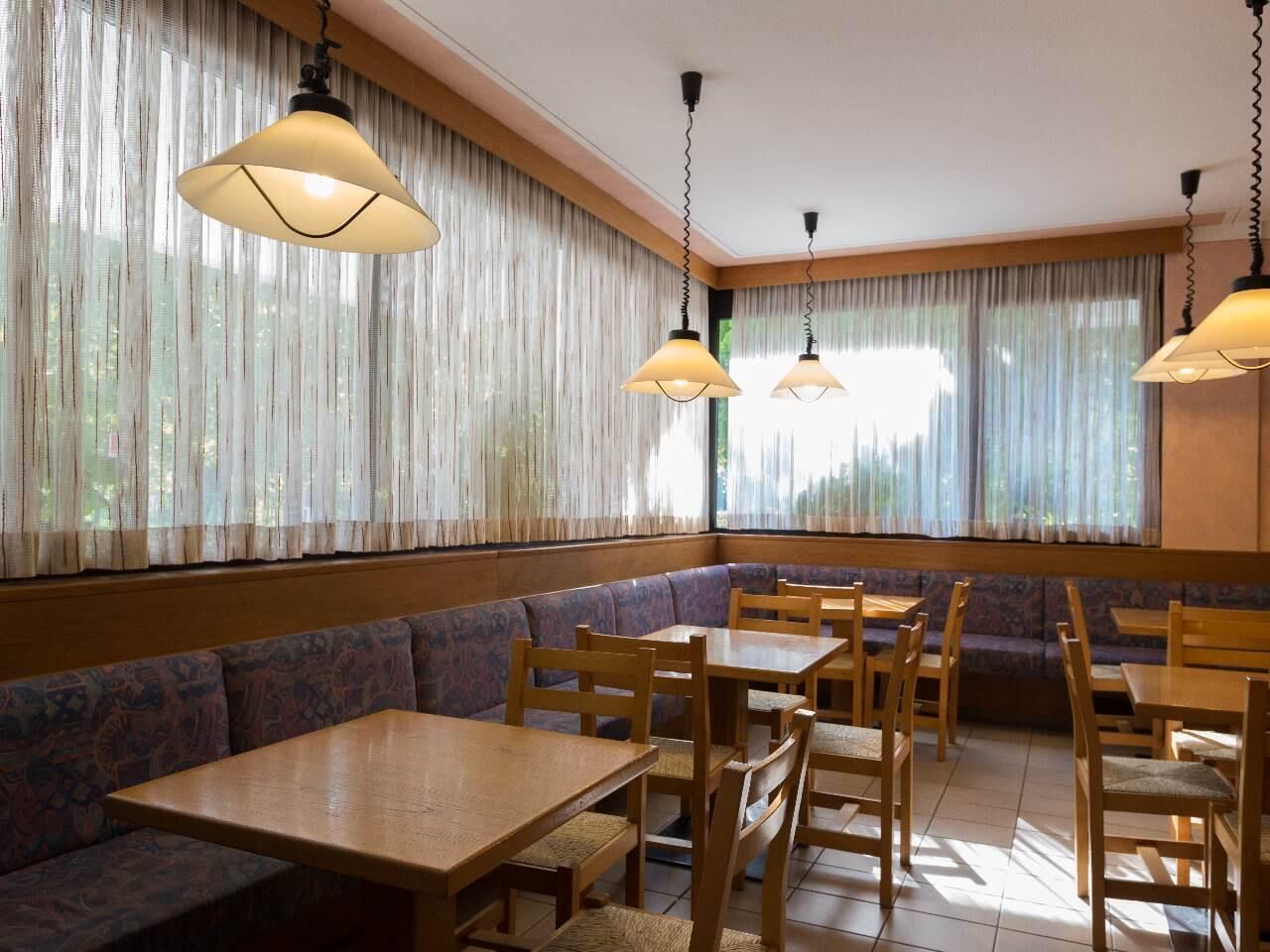 A restaurant with wooden tables and benches, hanging pendant lights, and large windows with sheer curtains.