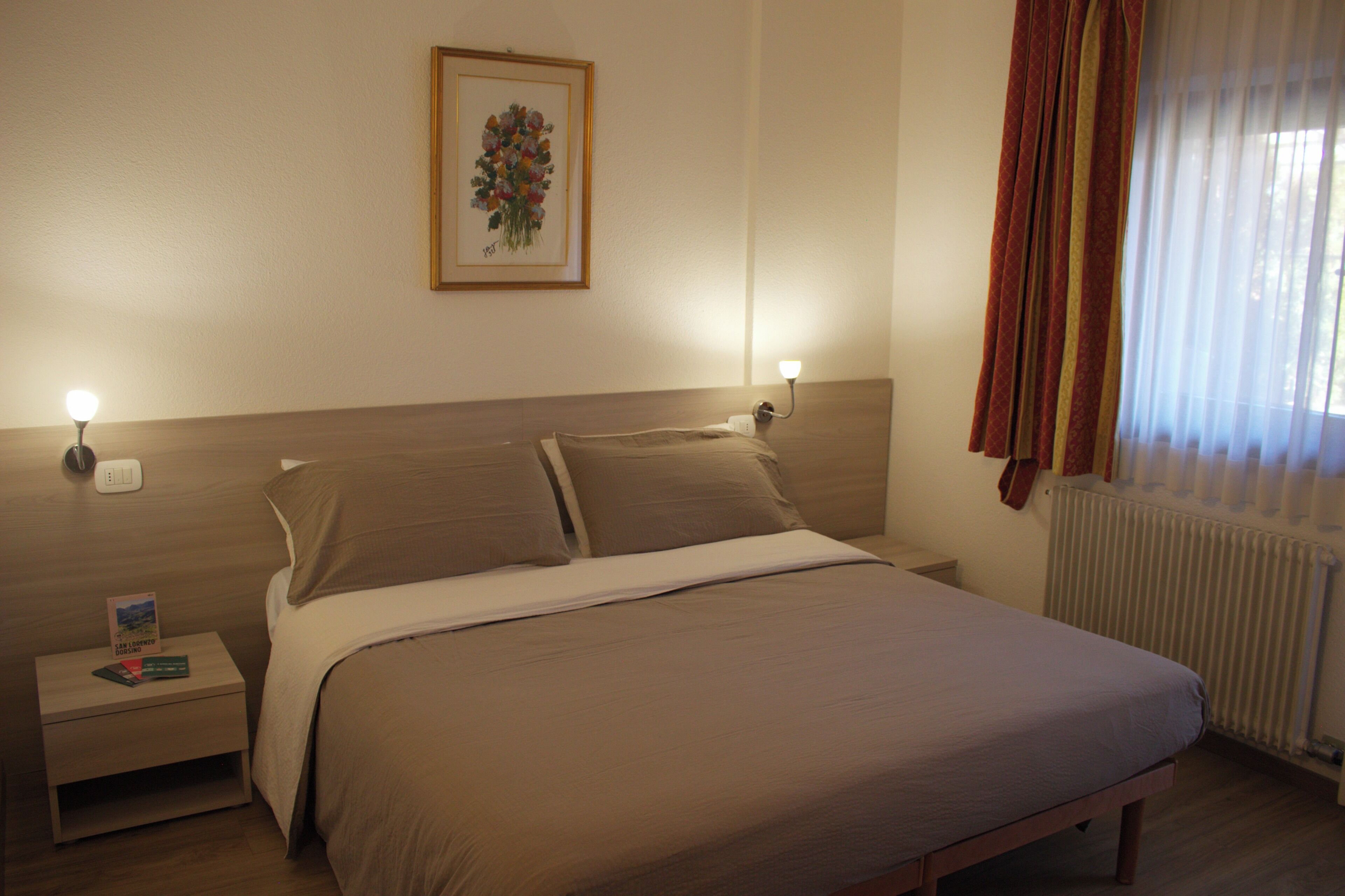 A bedroom with a bed, bedside tables, a nightstand, a framed picture on the wall, and a window with curtains.