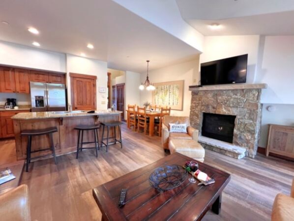 A spacious living area with a stone fireplace, wooden flooring, and a kitchen with wooden cabinets and a stainless steel refrigerator.