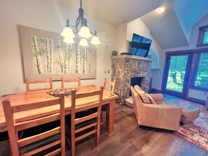 A dining area with a wooden table and chairs, a stone fireplace, and a large window with blinds.