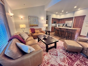 A living room with a sofa, coffee table, and a rug. There is a kitchen area with a refrigerator, microwave, and bar stools.