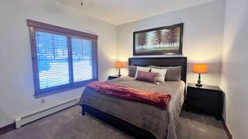 A bedroom with a bed, two bedside tables with lamps, a ceiling fan, a window with blinds, and a framed picture on the wall.