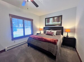 A bedroom with a bed, two bedside tables with lamps, a ceiling fan, a window with blinds, and a framed picture on the wall.