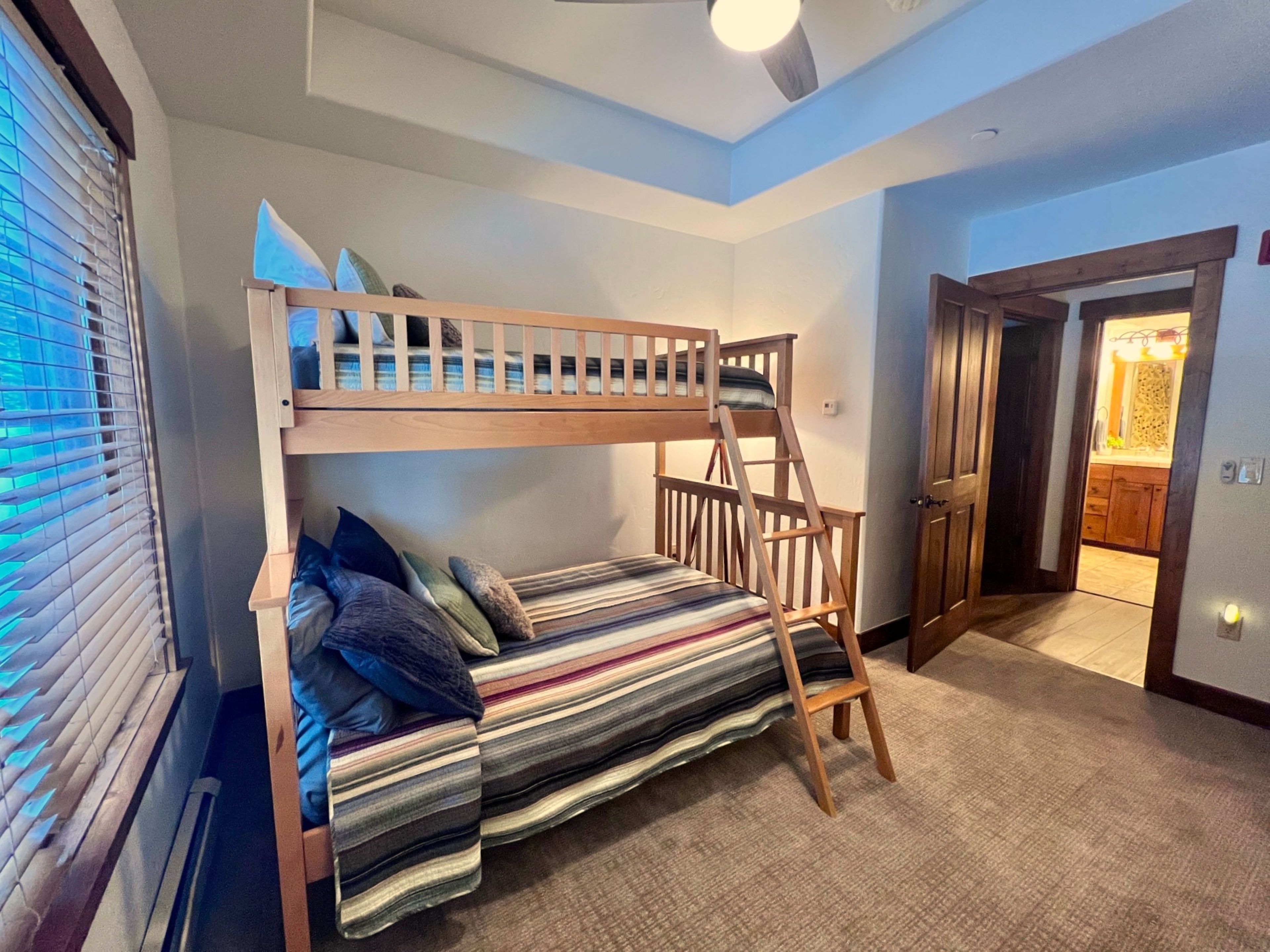 Una cama litera con escalera, una ventana con persianas y una puerta que da a un baño.