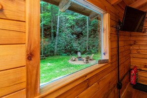 A wooden cabin with a large window overlooking a forested area. Inside, there is a wooden bench and a small table with chairs. - Cozy Creek | Private, Hiking, Creekside + Firepit (Rogers)