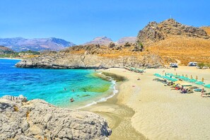 A beach with clear turquoise waters, rocky cliffs, and a sandy shore with umbrellas and beach chairs. - Villa Evelina | Private Pool (Adelianos Kampos)