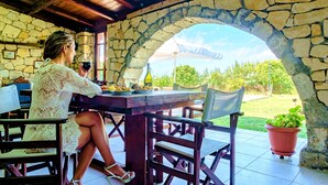 A stone archway leading to an outdoor patio with a table and chairs, a potted plant, and a bottle of wine. - Villa Evelina | Private Pool (Adelianos Kampos)