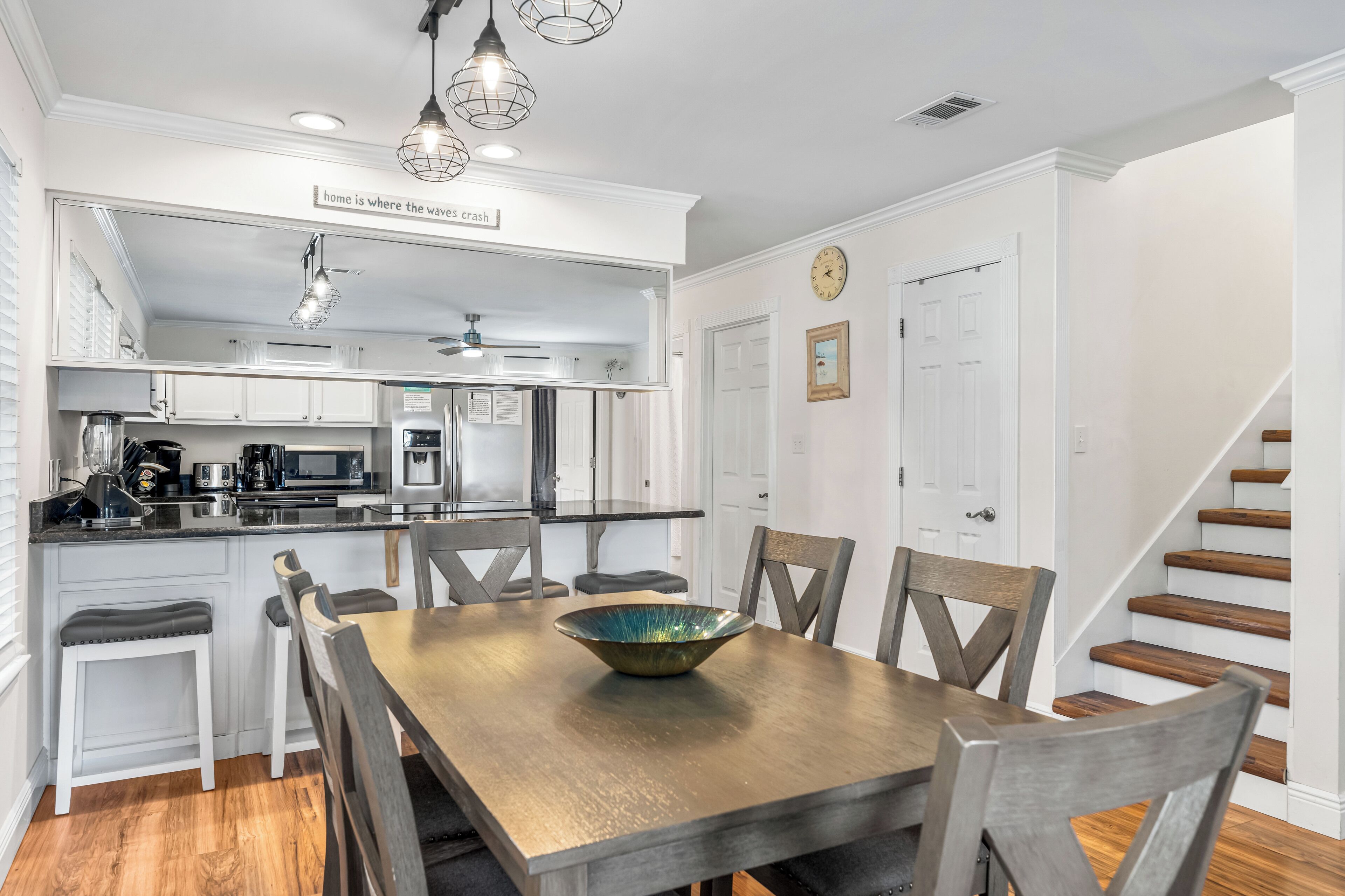 A kitchen with white cabinets, a wooden dining table with a bowl centerpiece, and a staircase to the right.