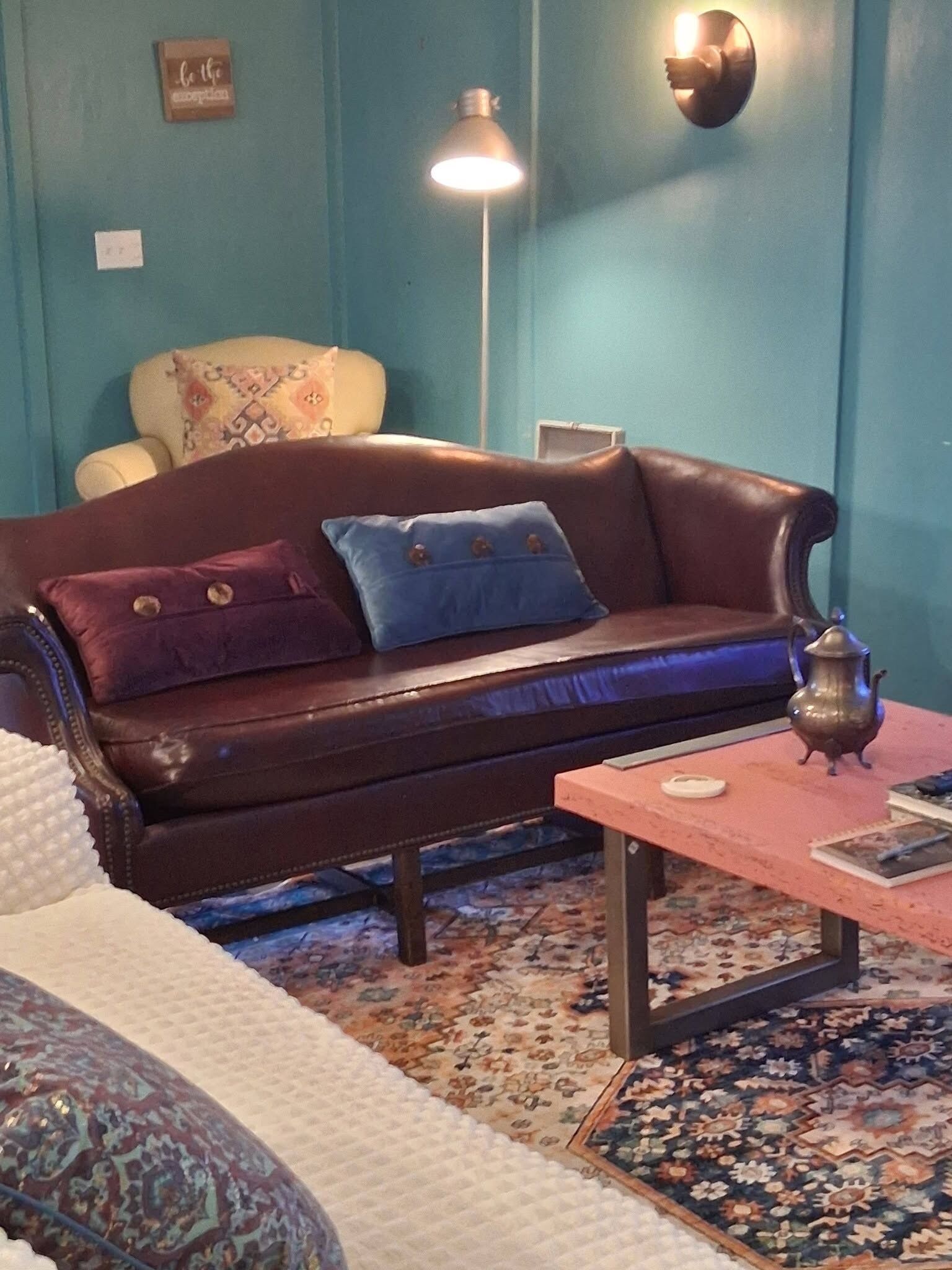 Una habitación con un sofá de cuero marrón, un tapete estampado, una mesita con tetera y una lámpara de pared.