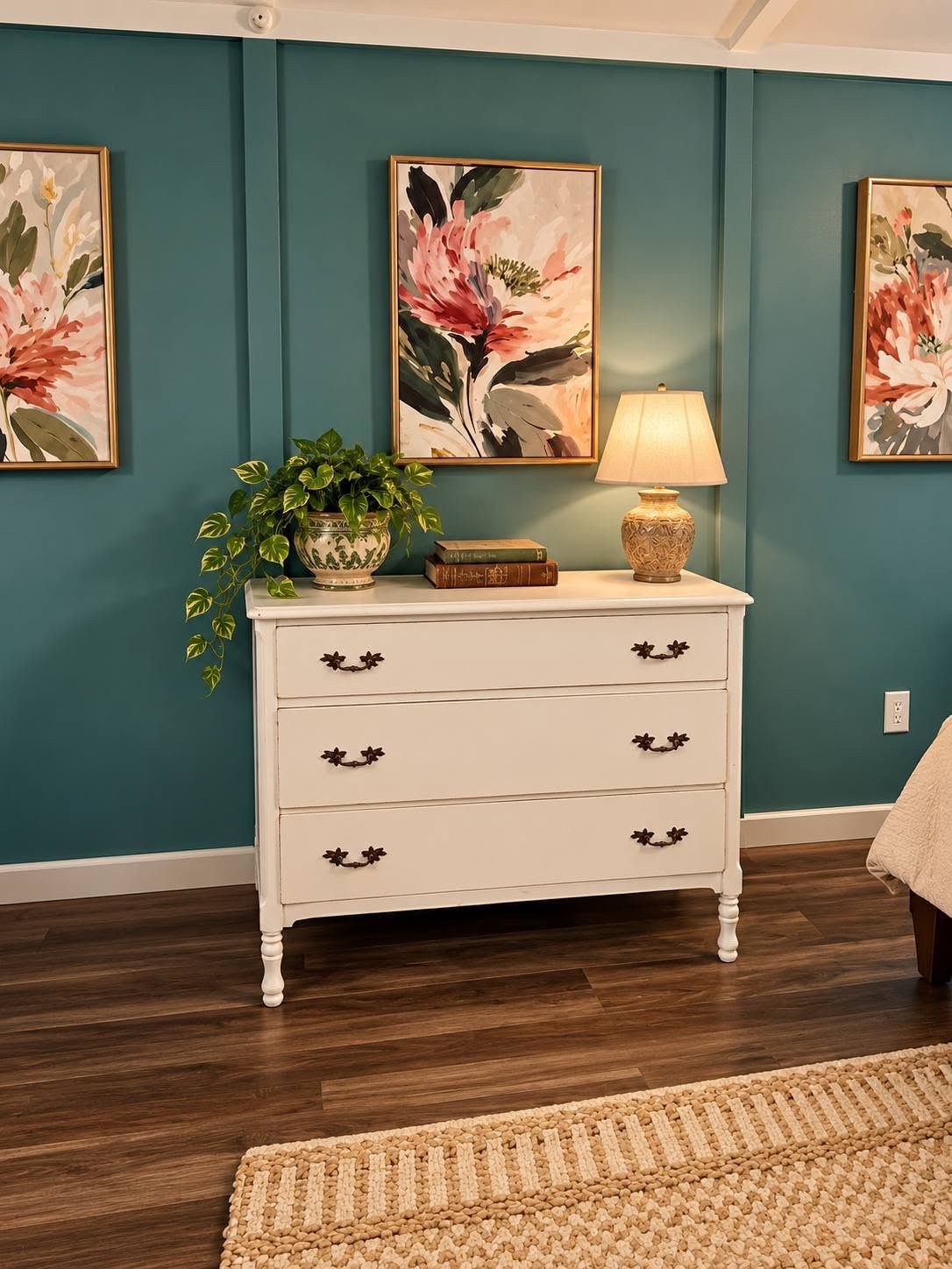 A room with teal walls, a white dresser with floral handles, a potted plant, and framed floral artwork.