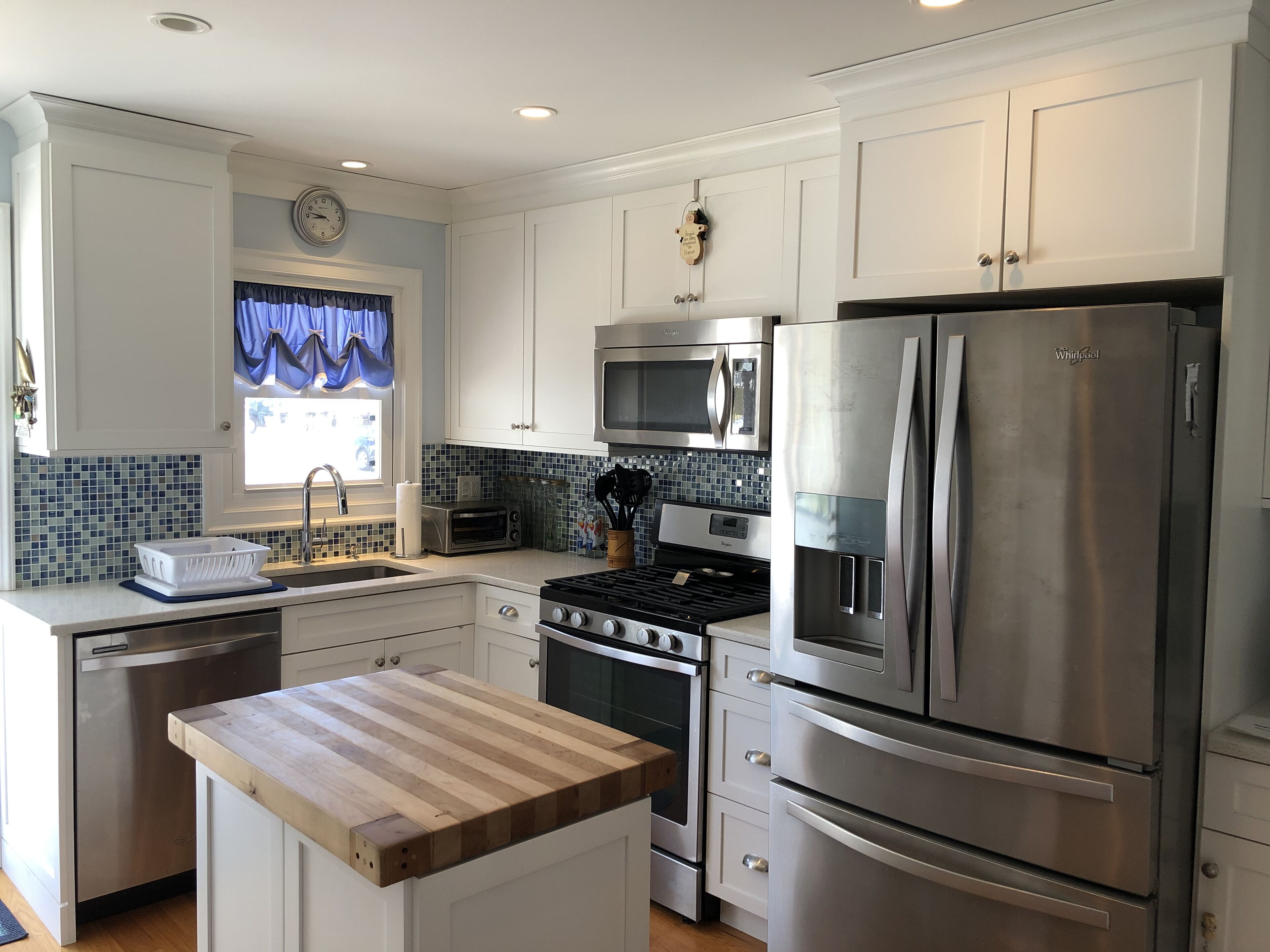 A modern kitchen with white cabinetry, stainless steel appliances, a wooden countertop, and a window with blue curtains.