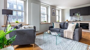 A modern living room with a grey sofa, a glass coffee table, and a patterned rug. There are large windows with blinds, a black floor lamp, and a kitchen area with dark cabinets and a microwave.