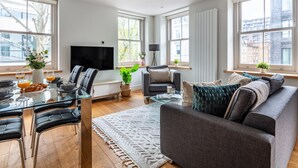 A modern living room with a glass dining table, black leather chairs, a grey sofa, and large windows offering city views.