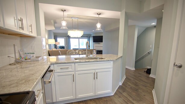 A modern kitchen with white cabinetry, a granite countertop, and a central island with a sink. There are pendant lights above the island and a staircase leading to another level. - Charming townhouse with beautiful ocean views and only steps to the beach! (Wrightsville Beach)