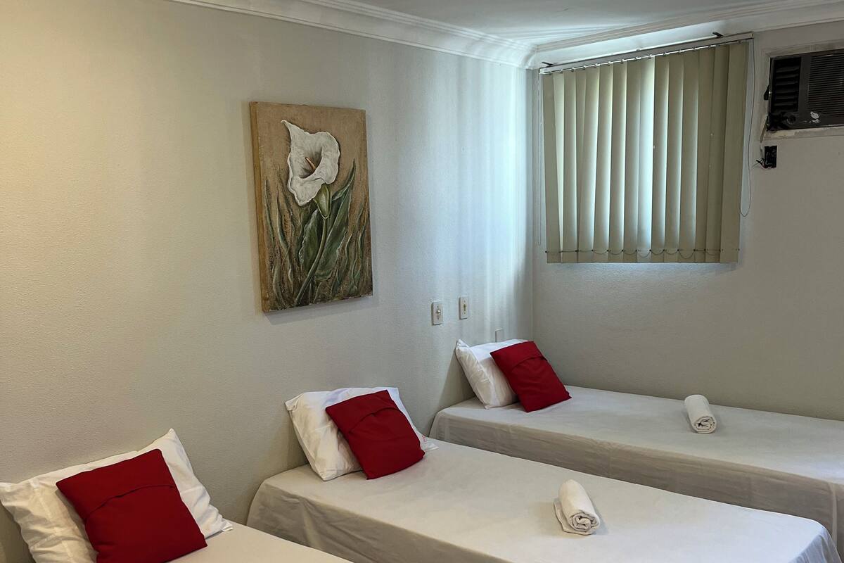 Quarto de hotel com duas camas de solteiro, cada uma com roupa de cama branca e almofadas vermelhas, um quadro de uma flor na parede e uma janela com persianas.