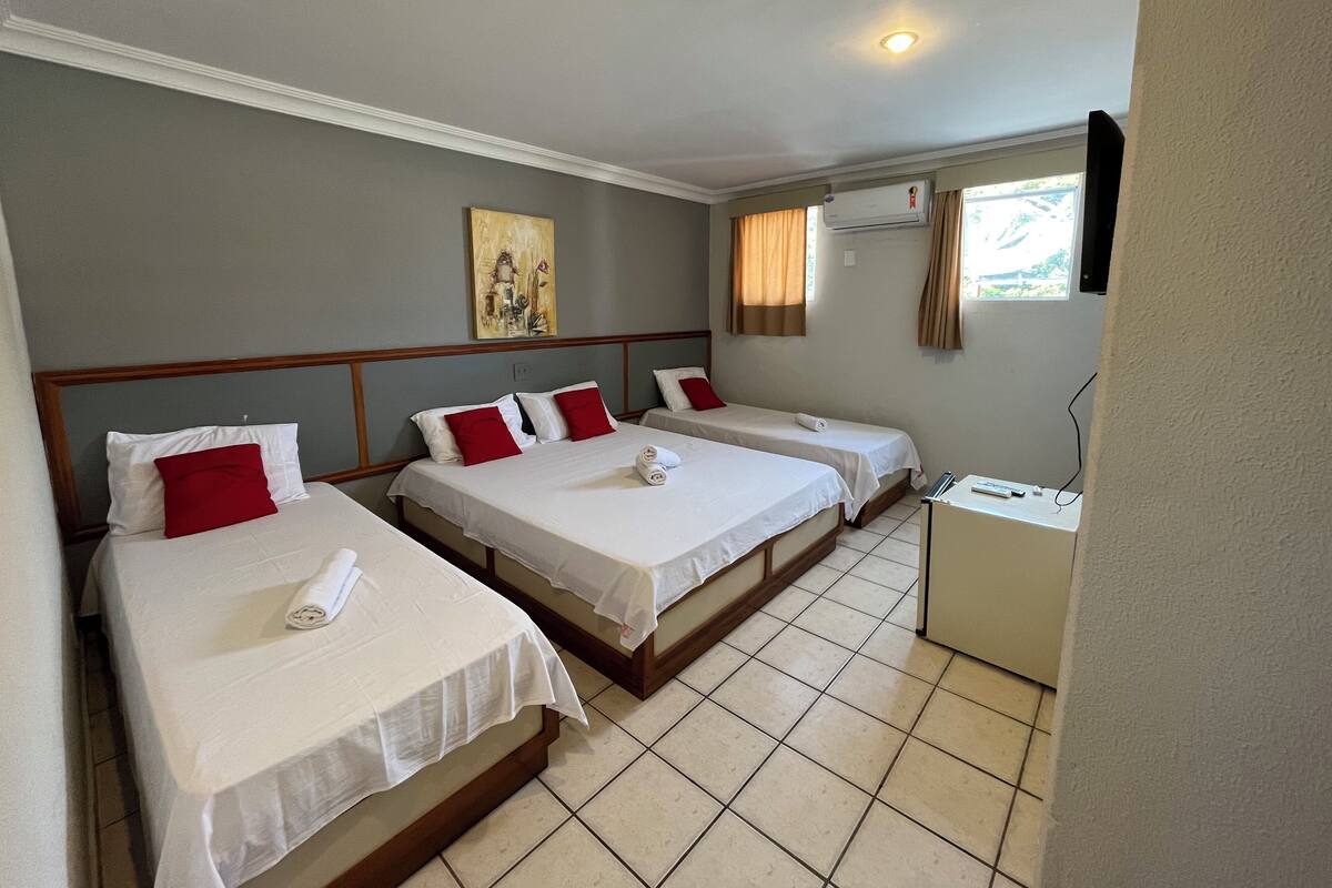 Quarto de hotel com três camas de solteiro, uma pequena escrivaninha, ar condicionado e um quadro na parede.