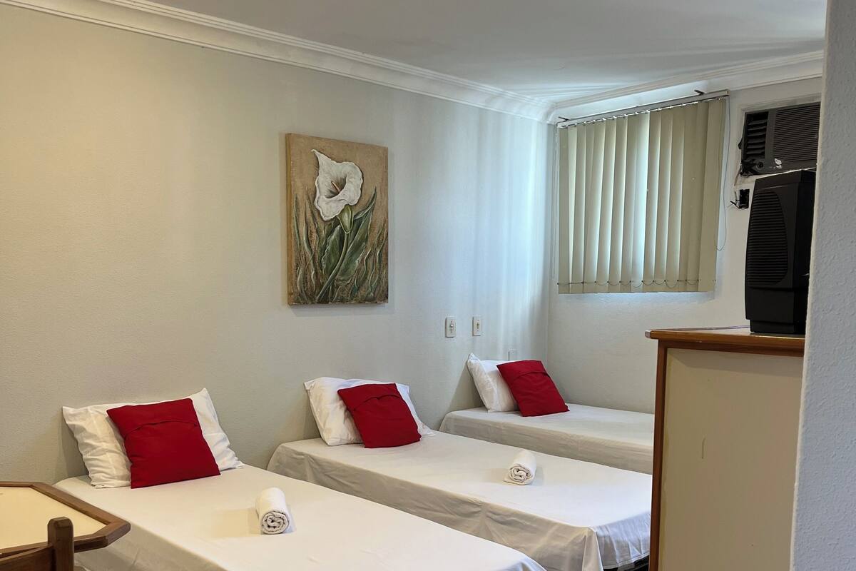Quarto de hotel com duas camas de solteiro, uma cadeira de madeira, uma mesa pequena e um quadro na parede.