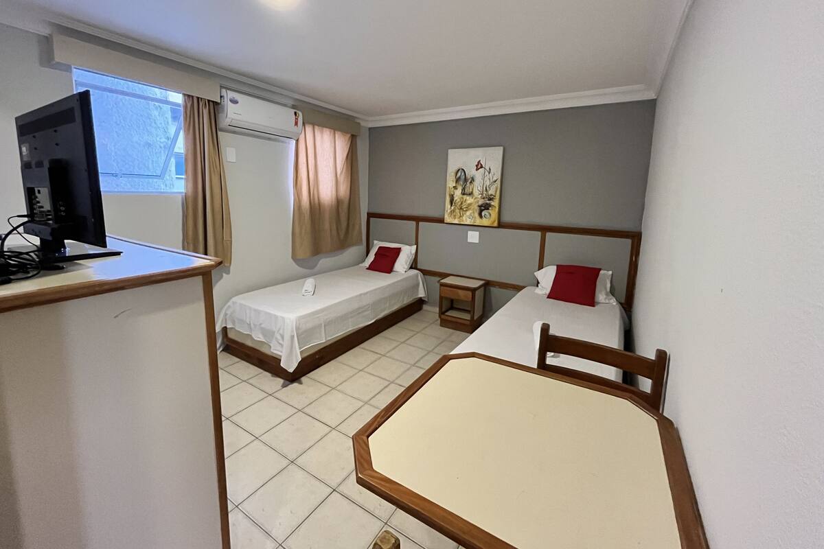 Quarto de hotel com duas camas de solteiro, uma escrivaninha com computador, uma mesinha e um quadro na parede.