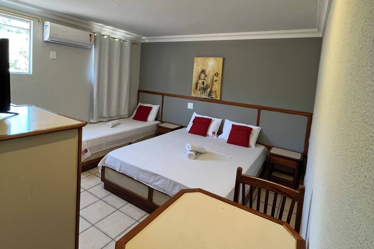 Quarto de hotel com duas camas, uma escrivaninha e uma cadeira. Há um ar condicionado na parede e um quadro sobre as camas.