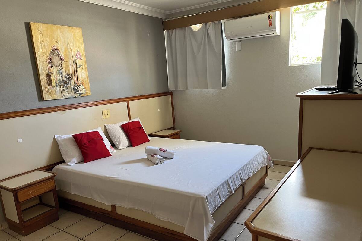 Quarto de hotel com cama, duas mesas de cabeceira, televisão e ar condicionado.