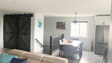 Una sala de estar moderna con un sofá color beige, un comedor con mesa y sillas, una cocina con refrigerador y un amplio ventanal que proporciona luz natural.