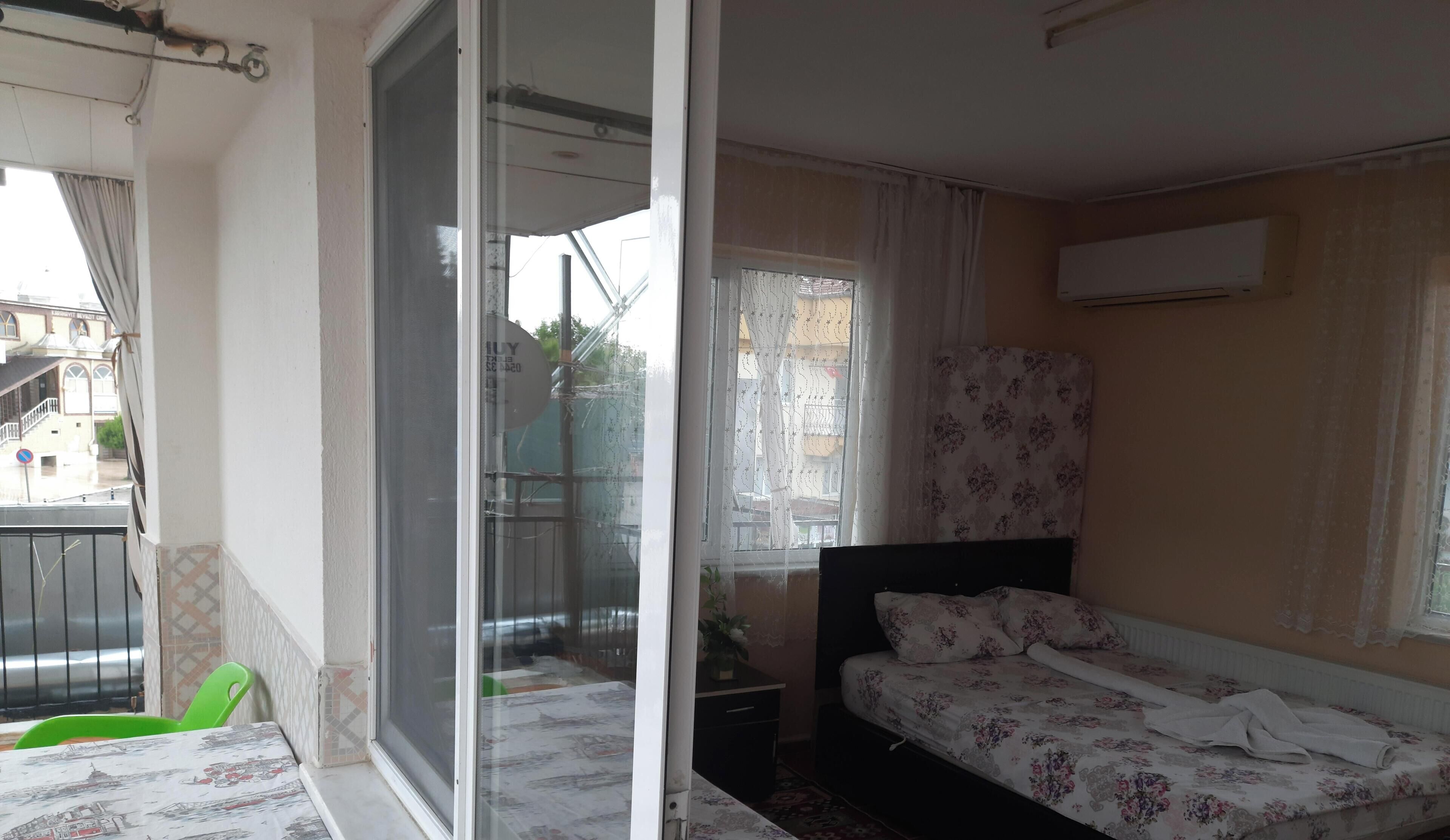 A bedroom with a bed, a window with curtains, a radiator, and a balcony with a chair.